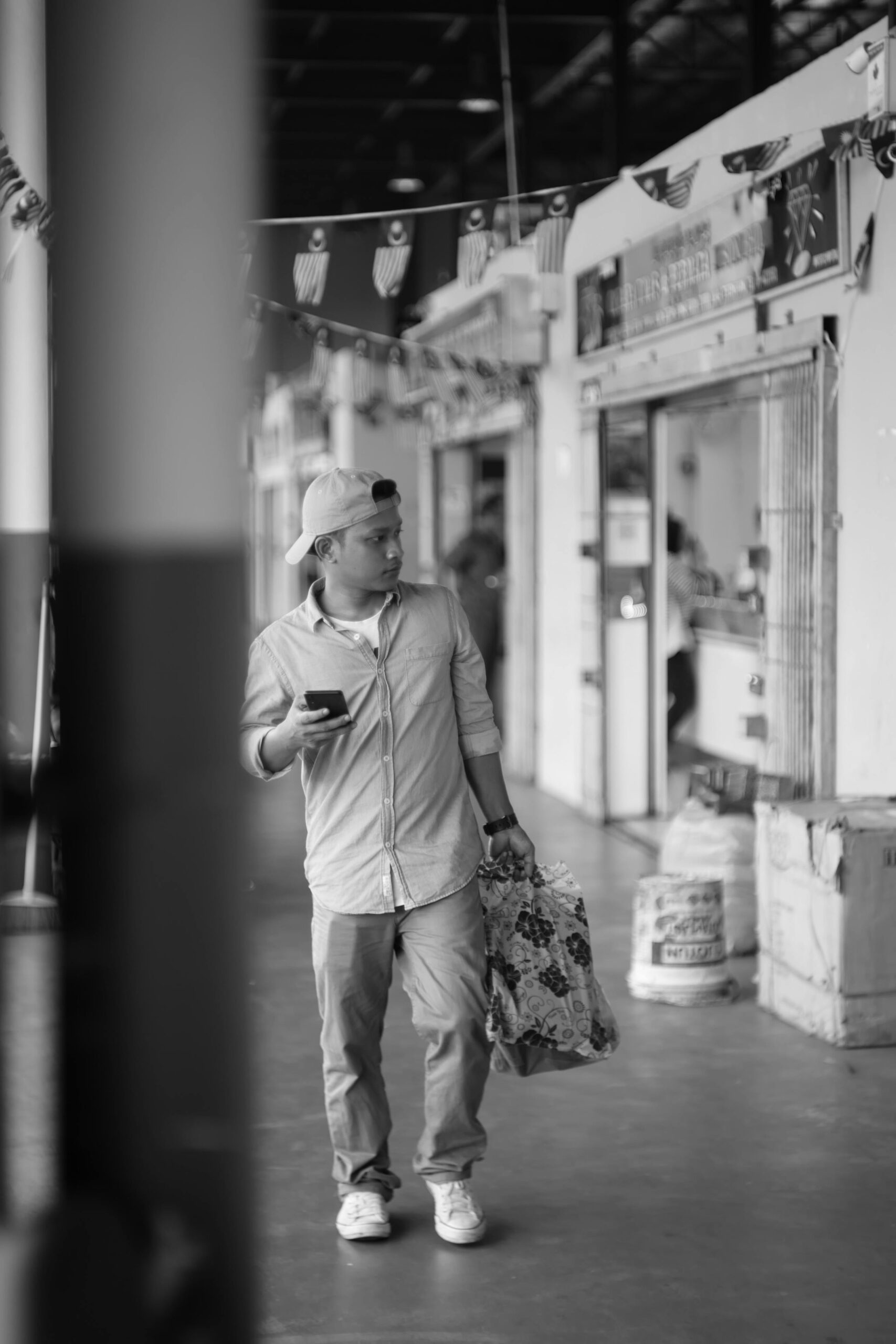 A man in casual attire walks through Kuala Terengganu market, holding a smartphone and bag.